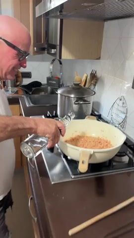 An older man is standing at a stove, pouring a clear liquid from a glass bottle into a pan of diced salmon. The pan is light cream in color with a light brown wooden handle, and is sitting on a black stovetop. Behind the stovetop is a dark brown counter, a stainless steel sink, a pot with a lid, and an arrangement of kitchen utensils. The man is wearing glasses and a white t-shirt. He appears to be filming a video.