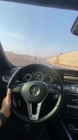 A person is driving a Mercedes-Benz on a paved road. The driver's right hand is visible on the steering wheel. The dashboard and windshield show the road ahead, featuring telephone poles, mountains, and a walled structure. The camera angle then shifts to show the driver, a man with a beard wearing a purple t-shirt and jeans. The setting appears to be during the daytime.
