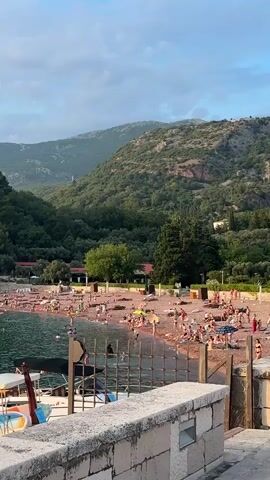 The video pans across a crowded beach with red-colored sand, situated at the base of lush, tree-covered mountains. Numerous people are visible on the beach, some sunbathing on towels and chairs, others wading or swimming in the clear blue water. A few boats are docked near the shore. The sky is partly cloudy, with a soft, diffused light suggesting either early morning or late afternoon. The scene conveys a sense of leisure and summer activity in a picturesque natural setting.