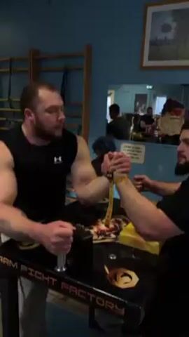 A muscular man with a beard and a black tank top is arm wrestling another man in a black shirt at a table with "ARM FIGHT FACTORY" printed on it. They are using a strap to secure their hands. The man in the black tank top has a tattoo on his left arm. There is a hand grip on the table for the arm wrestling.  In the background, there is a wall ladder, a mirror showing several people sitting at a table, and a framed picture on the wall.