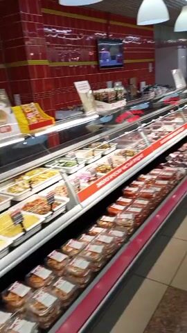 The camera pans across a deli counter inside of a supermarket. The deli counter features a wide array of salads, prepared foods, and meat products, all sealed in clear plastic containers. The counters are lit by overhead fluorescent lights. A woman wearing a white hat and a dark jacket walks into the frame and appears to be looking at the food. A sign on the counter reads "ЭКОЛОГИЧЕСКИ ЧИСТЫЙ ПРОДУКТ" in Russian, which translates to "ECOLOGICALLY CLEAN PRODUCT". The floor is tiled in a checkerboard pattern. A television screen is mounted on the red tile wall above the deli counter.
