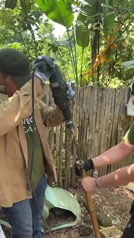 Two men work together outdoors in a wooded area with a bamboo fence in the background. One man, wearing jeans, a green t-shirt, and a tan jacket, holds an electric drill pointed downwards into the end of a bamboo pole held by the other man. The bamboo pole is held upright. The man holding the pole is wearing a green shirt, black pants, a black baseball cap, and black gloves. He has a beard. A large drill bit extends out of the drill. There are broken pieces of plastic on the ground.