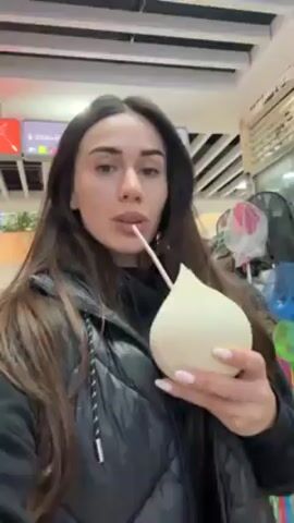 A woman is standing indoors, holding a white, teardrop-shaped container with a straw in it. She is drinking from the straw. She is wearing a black vest and has long brown hair. Behind her is a shop or stall with various items on display, including umbrellas and decorations. The background suggests she is in a public area, possibly an airport or shopping mall.