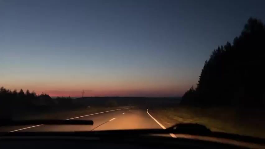 A first-person perspective from inside a car shows a road at dusk. The sky transitions from a dark blue at the top to shades of pink and orange near the horizon. The road curves to the right, with headlights illuminating the asphalt. Trees line the right side of the road. There are no other vehicles visible on the road.