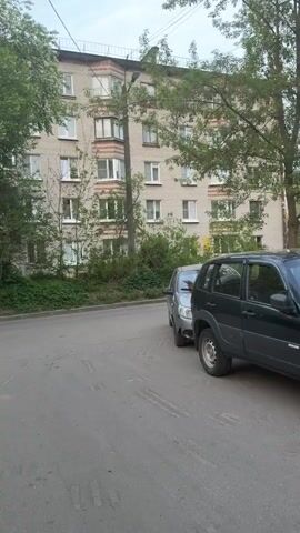 The video shows a person riding a dirt bike on a paved road.  The road is next to a multi-story brick apartment building, and there are several cars parked nearby.  There are trees and shrubs along the side of the road.  The person on the dirt bike is wearing a helmet and dark clothing.