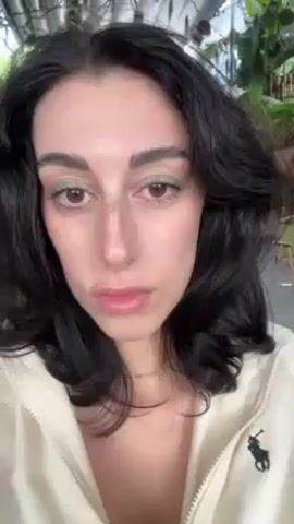 A woman is looking into the camera, her lips pursed slightly. She has dark, wavy hair that frames her face, and she's wearing a light-colored zip-up jacket with a Ralph Lauren logo on the left side. Her makeup includes a light green eyeshadow. The background appears to be an outdoor or semi-outdoor space, with greenery and what look like chairs visible.
