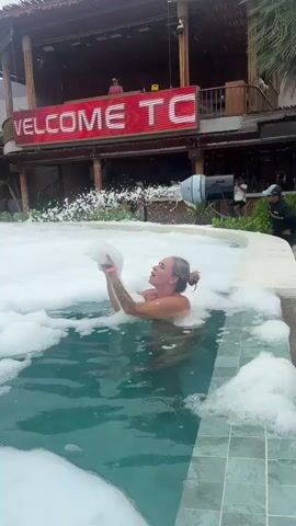 A woman is in a pool filled with white foam. She is holding a large clump of foam above her head and looking up. She has tattoos on her arms. Behind her, a building with a thatched roof is visible. A large red sign with white lettering that says "WELCOME TO 79" is mounted on the building. There are people in the background on a balcony and near the building. A large industrial fan is also visible to the right of the sign. The pool water is a greenish color. The overall scene appears to be an outdoor party or event.