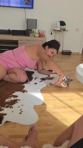 Here is a description of the video:

A woman in pink pajamas is on her knees on a cowhide rug, interacting with a cat. She gently pets and rolls the cat around, which appears playful. The scene takes place in a living room with a wooden floor. In the background, there's a TV on a cabinet, a small table with a lamp, and a doorway. A person is sitting on the floor in the foreground, with their legs visible.