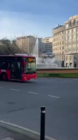 The video shows a street scene with vehicles passing by a roundabout with a fountain in the middle. 

The video begins with a red city bus driving past the roundabout. A black SUV follows, then a white van. Buildings of varying heights are visible in the background. The sky is blue with scattered clouds. The camera is positioned on the sidewalk, capturing the traffic flow and the urban landscape. The fountain in the roundabout is actively spraying water, adding movement to the scene.