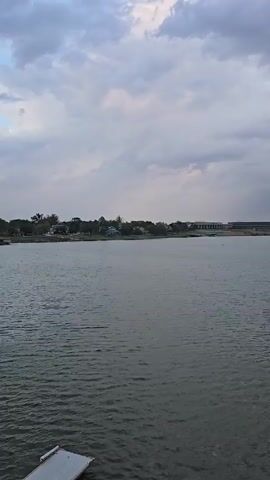A wide shot captures a body of water with a cloudy sky overhead. In the distance, a shoreline is visible with buildings and trees. The water has small ripples on its surface, suggesting a light breeze. The lighting indicates it may be early morning or late evening due to the soft, diffused light. No people or specific actions are clearly discernible.