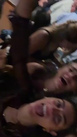 A close-up video captures three young women in a dimly lit, crowded indoor setting, possibly a party or club. The camera is handheld and shaky, focusing primarily on the faces of the two women in the foreground. They are laughing and appearing to be in good spirits, with their faces close together. One woman is on top of the other, with her arm around her. A third woman's face is visible behind them, also smiling. In the background, other people are blurred and out of focus, suggesting a lively atmosphere. The lighting is dim and possibly colored, typical of a nightlife venue.