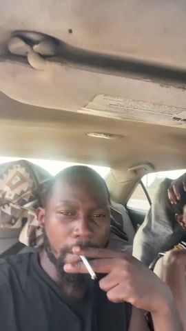 The video shows two men in the interior of a car, presumably during daytime. The man in the foreground is close to the camera, smoking a cigarette and looking directly at it. He has a beard and is wearing a black t-shirt. Behind him, to the right, another man is visible, partially obscured by a gray hooded garment draped over his shoulders. He is looking away from the camera, touching his head. The car's interior appears worn, with visible signs of use on the ceiling upholstery, which also has a label with some text. The background shows a blurry outdoor scene, suggesting they are in transit. The overall atmosphere seems casual, perhaps indicating a journey. The location context provided is "Immigration Quarters, Nigeria." There is no clear indication of the time of day or weather conditions beyond it appearing to be daylight.