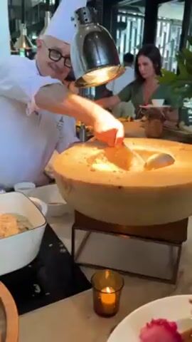 A chef in a white uniform and chef's hat uses a ladle to scoop risotto from a large wheel of cheese into a small white bowl. The cheese wheel sits on a wooden stand, illuminated by a heat lamp above. The chef wears glasses and is smiling. Several people are at a buffet behind him. The environment looks like a modern, upscale restaurant with large windows and green plants. A lit candle is on the counter in front of the cheese wheel.