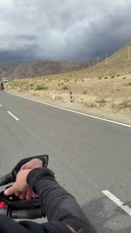 The video begins with a close-up shot of a person's arm, wearing a black compression sleeve, extending a thumb up towards the camera. The arm is positioned in the foreground, obscuring part of the view. The background shows a paved road with a white line marking the edge. To the right of the road, a dry, arid landscape with sparse vegetation and rocky hills stretches towards a cloudy sky. A few utility poles are visible on the hillside.

The camera then pans slightly, revealing a motorcycle's side mirror on the left. Shadows of two people on a motorcycle are cast on the road.

The scene shifts to a wide shot of the road and the surrounding landscape. A child, wearing a yellow t-shirt, blue shorts, a black helmet, and sunglasses, is riding a red bicycle. The bicycle has a black bag attached to the rear rack. The child appears to be being towed by a rope connected to the handlebars, which disappears out of frame to the right.

The view returns to the perspective of someone on a motorcycle. A white van with a yellow license plate approaches from the opposite direction on the road and passes by. The sky is overcast with dark clouds. The shadows of the two people on the motorcycle are again visible on the road. The video ends with the van moving away in the distance.