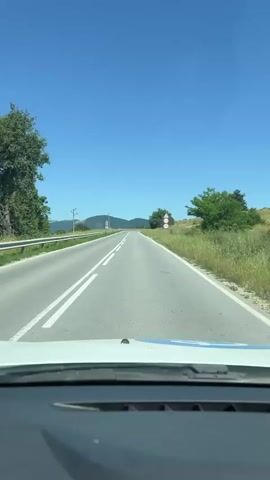 The video shows a view from the driver's seat of a car moving along a paved road. The road has two lanes separated by a double white line and a single white line on the right edge. The sky is clear blue. On the right side of the road, there is a grassy slope with a few trees and a sign with a speed limit of 70. On the left side of the road, there is a guardrail and more grassy slopes with scattered trees and a utility pole. In the distance, rolling hills are visible. The car's dashboard is visible at the bottom of the frame.