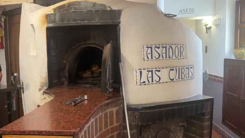 A traditional, large wood-fired oven is the central focus of this video, likely situated within a restaurant in Arévalo, Spain. The oven features a white, plastered exterior over a brick base, with a red granite countertop in front. Inside its dark, arched opening, food – possibly poultry or meat – can be seen cooking in a pan. A long, metal, oven-tending tool leans against the oven's side, and a pair of metal tongs rests on the countertop. On the white facade of the oven, decorative blue and white tiled letters spell out "ASADOR" (roasting oven/restaurant) and "LAS CUBAS", identifying the establishment as "Asador Las Cubas". A small sign above the oven entrance reads "ASEOS" (restrooms). The camera slowly zooms in on the oven throughout the video, highlighting its rustic and functional aesthetic, indicative of a traditional Spanish eatery.