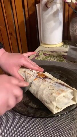 A close-up shot shows a person cutting a wrap in half on a dark-colored plate. The wrap is rectangular and filled with ingredients, including what appears to be orange carrots and shredded purple cabbage. The person uses a silver knife with a black handle to saw through the wrap, their left hand holding the wrap in place. The filling of the wrap is visible where it's cut. The plate rests on a grey and white patterned placemat. In the background is a wooden wall, a paper towel holder with a roll of paper towels, and a vase. The lighting is slightly warm.