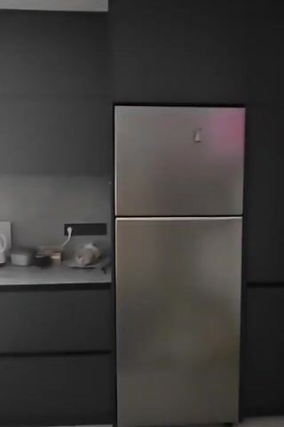 The video pans across a modern kitchen with dark gray cabinets and stainless steel appliances. On the countertop, near the refrigerator, are two electric kettles, a small cup, and some packaged food items. To the left, a stove is visible, and a sink with a faucet and drying rack is situated near a window. Various items are on the windowsill, including bottles of oil and cleaning supplies. The overall impression is a functional and tidy kitchen space.