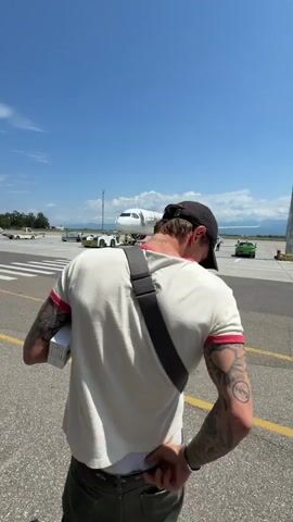 The video shows a man walking on an airport tarmac. He is wearing a black baseball cap, a white t-shirt with red trim on the sleeves, green pants, and a crossbody bag. He has tattoos on his arm and is wearing a watch. He turns to the camera and makes a peace sign with his fingers.

In the background, an airplane with pink accents is visible, as well as a jet bridge, airport vehicles, and other people walking towards the plane. A bright green car is parked in the distance. The sky is clear and blue, suggesting a sunny day. The overall scene depicts travel and the excitement of boarding an airplane.

Visible text:
- A "LA" logo is on the man's cap.
- There are tattoos on the man's arm with legible text, including "Love is..." and "BFF."