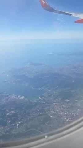 An aerial view from an airplane window shows a coastline with scattered buildings and inlets of blue water. The wing of the airplane, with an orange and white logo that reads "jet", is visible in the upper right corner of the frame. The sky is clear and blue.