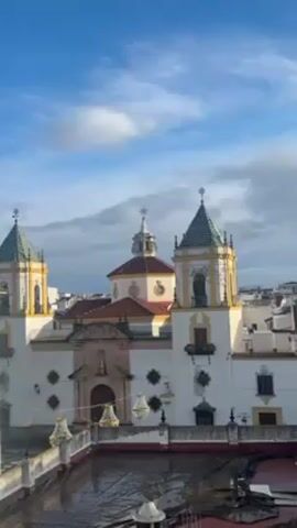 The video shows a view of a white and yellow church with two towers, a dome, and a cross on top of each. The sky is blue with some clouds. The church is in the middle of a city with white buildings. There is a red roof below the camera. The sound of church bells can be heard.