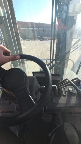 The video shows the inside of a forklift. The steering wheel, levers, and pedals are visible. A bottle of water sits to the right of the levers. Through the front and side windows, we see a gravel-covered yard with buildings, stacked wood, and vehicles. The camera pans slightly to show more of the yard. In the rearview mirror, a person's head is visible. The weather appears to be sunny.