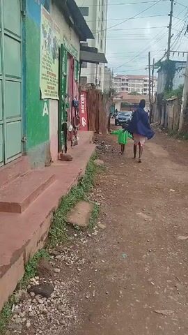 Here is a description of the video:
The video depicts an adult and a young child walking away from the camera on a narrow dirt road lined with buildings and fences. The adult, wearing a shawl and shorts, walks alongside the child, who is wearing a bright green jacket. The road surface is uneven, with dirt, rocks, and scattered debris. In the background, a car is parked, and other people can be seen walking further down the road. Buildings of varying heights flank the road. The overall impression is that of a residential area in a developing country.