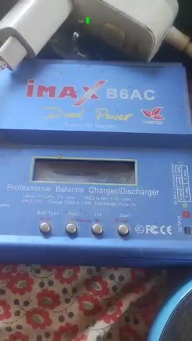 The video shows a close-up view of a blue IMAX B6AC charger. The charger has a small LCD screen and several buttons labeled "Batt. Type," "Start," "Inc.," and "Dec." There are also labels indicating different battery types and charge rates. To the right of the charger, there is a diagram showing battery connectors. The charger is placed on a surface covered with a red and white floral patterned cloth. In the background, a white power adapter and some cables are visible. To the left of the charger, a roll of black tape and a small cardboard package with a yellow price tag are present. The overall lighting is dim.