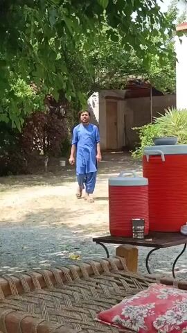 The video depicts a man in a blue shalwar kameez walking on a gravel pathway in a yard. He's walking towards the camera. In the foreground, there is a traditional woven cot with a red patterned pillow on it. A small metal table next to the cot holds two red water coolers and a small metal cup. The yard is surrounded by green trees and shrubs. In the background, a white building is visible. The sky is bright and slightly overcast.