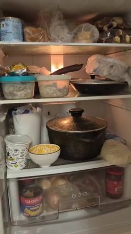 The video pans across the interior of an open refrigerator. The shelves are filled with various food items, including containers of prepared food, packaged goods, and a large pot. A light illuminates the interior of the refrigerator.