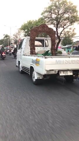 A white Mitsubishi pickup truck is driving down a paved road. The truck bed contains a large, decorative wooden archway and a green tarp. The truck has a license plate that reads "H 8131 PM". The truck is in the middle lane of traffic, and other vehicles, including motorcycles and a van, are visible on the road. Trees and buildings are present in the background. The overall scene appears to be an urban or suburban street during daylight.