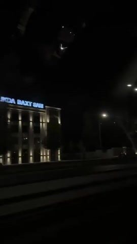 This video captures a nighttime view from inside a vehicle, likely moving along a road. The scene is dominated by darkness, with artificial lights providing the primary illumination.

On the left side of the frame, a building with illuminated signage is visible. The signage appears to read "CK SARI" in blue letters against a lighter background. The building itself is modern in design and is lit by spotlights. Adjacent to the building, a low wall or barrier is illuminated by a series of evenly spaced golden lights. Trees are sparsely planted behind this illuminated barrier.

The road surface is dark and appears to have multiple lanes, indicated by contrasting horizontal lines. Streetlights, also featuring bright, modern fixtures, are positioned along the road.

The overall impression is of a developed urban or suburban environment at night, characterized by functional and decorative lighting. There are no people clearly visible within the frame, and the focus is on the architectural and environmental lighting elements. The camera appears to be panning slightly or the vehicle is in motion, causing a subtle shift in the perspective.