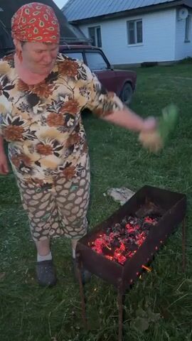 A woman is standing outdoors next to a barbecue grill with hot coals inside. She is fanning the coals with a large, flat object, apparently a large leaf or piece of cardboard. The woman is wearing a red bandana, a floral shirt, patterned shorts, and clogs. The setting appears to be a backyard with a white house and a maroon car visible in the background. The grass is green, and the lighting indicates it is dusk or evening. The woman is smiling.