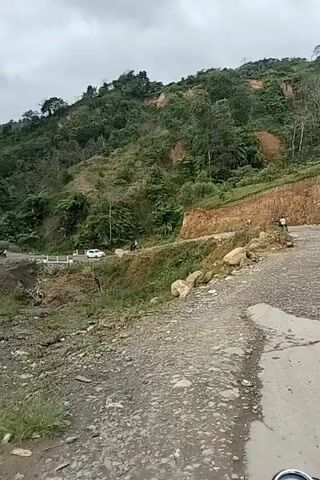 The video shows a mountainous landscape with a dirt road winding through it. Lush green vegetation covers the hillsides, but there are also areas of exposed earth and rocks, indicating potential erosion or landslides. A white car is visible on the road in the distance, as is a person walking near it. The road appears to be rough and uneven, with loose gravel and rocks. The sky is overcast, suggesting cloudy weather. The camera appears to be mounted on a moving vehicle, possibly a motorcycle, given the slight vibration and perspective.