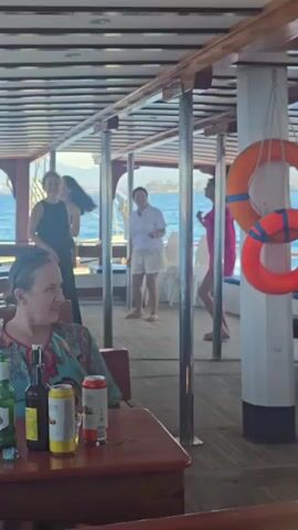 A group of women are on the deck of a boat. In the foreground, a woman in a brightly colored tunic sits at a table with several bottles and cans of drinks. In the background, three women are standing. One woman in a black dress is talking on a microphone, while another woman in a white shirt and shorts stands nearby. A third woman in a pink dress is looking away from the camera. The boat is on the water, with land visible in the distance. The deck is made of wood, and there are railings and support beams. Orange life preservers hang on the right side of the frame. The overall atmosphere appears to be casual and celebratory.