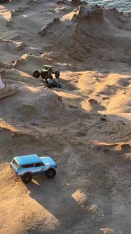 The video shows three remote-controlled vehicles on a sandy, rocky terrain. A blue SUV sits in the foreground. A green monster truck is higher up on the terrain, and a blue buggy is even further uphill. One person's leg and foot are visible in the lower left corner of the frame. The lighting suggests late afternoon or early morning. In the background, a body of water is visible. The blue buggy is climbing uphill.