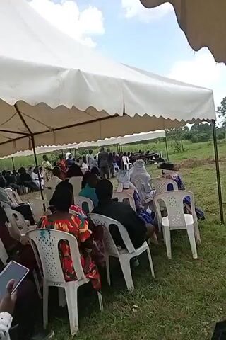 A group of people are gathered under a large white tent that is erected in a grassy field. The people are seated in white plastic chairs and appear to be listening to someone speaking in the distance, off camera. The speaker’s voice can be heard in the audio. A person wearing a red headscarf and a white polka-dot shirt walks towards the front of the group. The sky is partly cloudy.