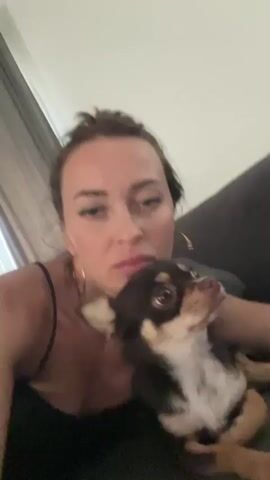 The video shows a close-up of a woman and a dog. The woman is wearing a black tank top and has her hair pulled up in a bun. She is wearing large hoop earrings. The dog is a small breed, possibly a chihuahua, with brown, black, and white markings. The dog is licking the woman's face, and the woman appears to be kissing the dog back. They are indoors, with a couch and a window with curtains visible in the background. The lighting is dim.