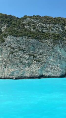 The video shows a boat moving through incredibly clear, turquoise water, approaching a dramatic coastline. Towering white limestone cliffs dominate the scene, covered in patches of green vegetation. The cliffs feature several large caves and indentations. As the boat continues, the view opens up to reveal a secluded beach nestled between two massive cliff faces. A shipwreck is visible on the sandy shore, along with several small boats and a few people in the water. The sky is a clear, bright blue.
