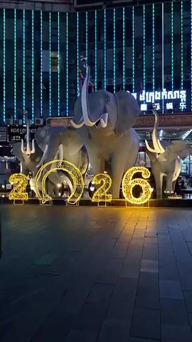 This video captures a nighttime scene in Sihanoukville, Cambodia, featuring a festive display of illuminated elephant sculptures and the numbers "2026" crafted from bright yellow lights. The elephants, of varying sizes, are positioned prominently in front of a building adorned with vertical, colorful LED lights that create a dynamic, pulsating backdrop. The largest elephant, centrally located, stands with its trunk raised, exuding a sense of grandeur. Smaller elephants flank it, adding to the procession-like feel of the display.

The "2026" numerals are large and bold, also illuminated with warm yellow lights, suggesting a celebration or commemoration, likely for the upcoming year. The ground is paved with dark tiles, reflecting the vibrant lights and adding depth to the scene. In the foreground, the rear of a dark-colored vehicle, possibly an SUV, is visible on the left, with its taillights showing a colorful reflection.

The overall atmosphere is one of cheerful festivity and perhaps anticipation, enhanced by the bright, artificial lighting against the dark night sky. The vertical streaks of light on the building's facade, ranging from white to pink, blue, green, and yellow, create a modern and energetic ambiance. The presence of text in what appears to be Khmer script on the building behind the elephants adds a specific cultural context to the location. The time of day is clearly night, given the darkness and the reliance on artificial illumination. There is no indication of weather conditions.