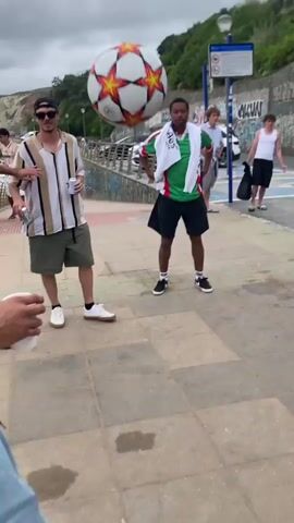 In an outdoor, paved area with a slight incline, several people are gathered. In the foreground, a person is partially visible from the waist down, wearing denim shorts and white sneakers with blue accents, their legs bent as if preparing to kick. Behind them, two men stand. One, on the left, wears a tan and white striped short-sleeved shirt, olive green shorts, a black baseball cap, and white sneakers, holding a white cup in his left hand. The other man, in the center, wears a green and white striped shirt, black shorts, and black sneakers, with a white towel draped over his shoulders. He is looking upwards, and a soccer ball with orange and white panels is in the air above him.

In the background, a white bus labeled "CITARO" is parked. To the right of the bus, a woman in a white top and black skirt walks away from the camera. Further in the background, more people are visible near a wall covered in graffiti. The environment appears to be a promenade or walkway with a view of a hillside and a cloudy sky. The individuals are engaged in playing with a soccer ball, with one person appearing to be kicking it or about to kick it, and another man focused on the airborne ball.
