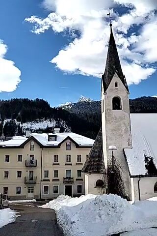 The video shows a white church with a tall steeple in a snow-covered village. The church has a dark roof and the main body of the building is covered in a blanket of snow. In the background, there are snow-covered mountains and a clear blue sky with some clouds. There are other buildings visible in the background, as well as some trees. The ground is covered in snow. In the last shot, a person is seen walking down the road, and several cars are parked.