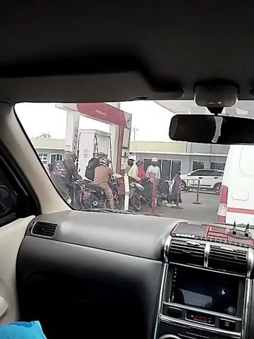 The video shows a view from inside a car looking out at a gas station. There are several motorcycles at the gas pumps, with people filling them up with fuel. The gas station appears to be busy, with a few other vehicles visible in the background. The weather is overcast.