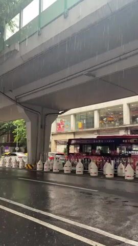 The video shows a street scene during a rain shower. The camera is positioned under an elevated structure, likely an overpass or bridge, which shelters the view from the direct rain. Heavy raindrops are visible falling throughout the frame. In the background, a red bus is stopped at a bus stop, with several people visible inside. The bus stop area is lined with white and red barrier cones. Beyond the bus stop, there are buildings with multiple windows, some of which appear to be shops or restaurants, with illuminated signs. The street is wet and reflects the light from the surroundings, indicating it is actively raining. A person holding a pink umbrella can be seen in the right side of the frame, moving away from the camera. The overall atmosphere is urban and wet due to the rain.