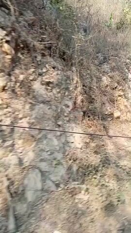 A handheld, first-person perspective video shows a rocky hillside with sparse vegetation. In the foreground, a pile of gravel is visible. A black pole with a red and white stripe is in the right foreground. The camera pans to the left, revealing more of the rocky terrain and a blue sky. A thin black cable is visible running horizontally across the frame in the middle ground.