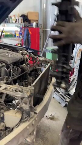 The video shows a man wearing a dark green baseball cap, a dark grey work jumpsuit, and black gloves. He is working on a car in what appears to be a garage or auto repair shop. The car's hood is open, revealing the engine and various components. The front of the car is partially disassembled. The man is holding a large rectangular object with a silver radiator-like core and black plastic frame. He carefully positions it in front of the engine, apparently fitting it into the car's frame. Tools and other objects are visible on shelves in the background.