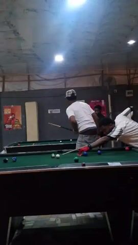 In a dimly lit indoor pool hall with a cracked, stucco-like ceiling, two men are playing pool. One man, wearing a white t-shirt, grey pants, and a patterned hat, stands by the pool table with a cue stick in hand. He glances at his companion who is wearing a black and white striped soccer jersey and red gloves. The soccer jersey-clad man bends low over the pool table to take a shot, then straightens up to observe the outcome. Other pool tables stand in the background, along with advertisements for drinks.