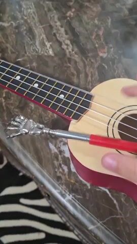 The video shows a close-up view of a ukulele on a marble-like surface. A person's hand is visible, plucking the strings of the ukulele. Also resting on the ukulele is a metal object that appears to be a retractable pointer or antenna, with a silver, skeletal hand-shaped end. The pointer has a red handle. The background shows a blurry pattern of black and white stripes, possibly fabric. The lighting suggests an indoor setting.