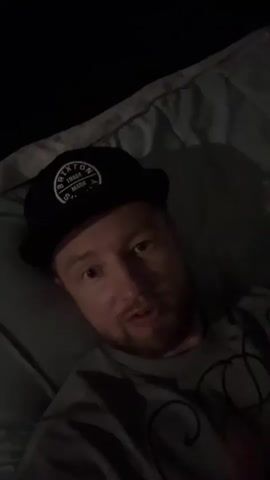 A man is lying on a bed and speaking to the camera. He is wearing a black baseball cap and a grey t-shirt. The room is dimly lit. He holds up two fingers in a peace sign gesture and then moves his hand. He smiles at the camera and waves.