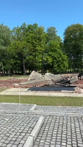 The video shows the ruins of a crematorium at a concentration camp. The ruins consist of broken concrete slabs and brick rubble. The ruins are surrounded by grass and a line of trees. In the foreground, there is a cobblestone pavement. The sky is blue and clear. The video pans from left to right, showing more of the ruins and the surrounding area. There's also white rope strung between short white poles.